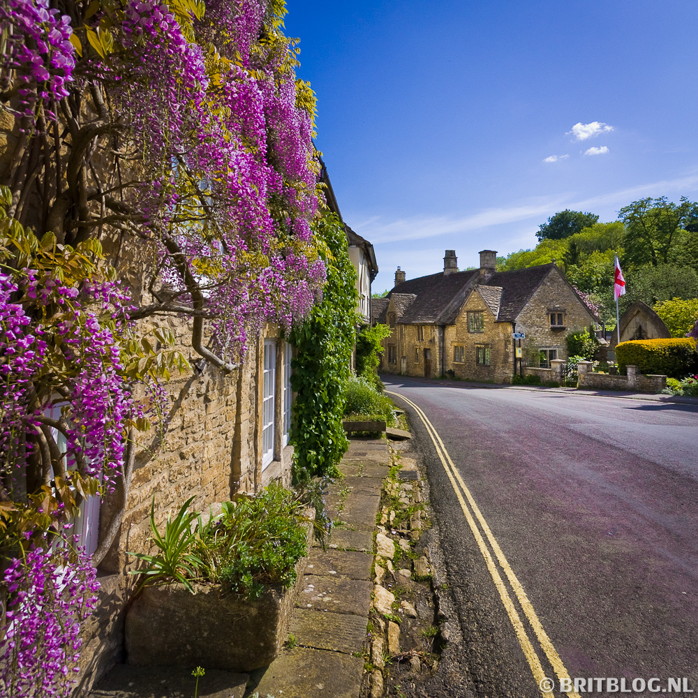 Kleurrijke straat in de Castle Combe, Cotswolds, met bloeiende paarse blauweregen tegen een stenen huis en een kronkelende weg richting charmante cottages