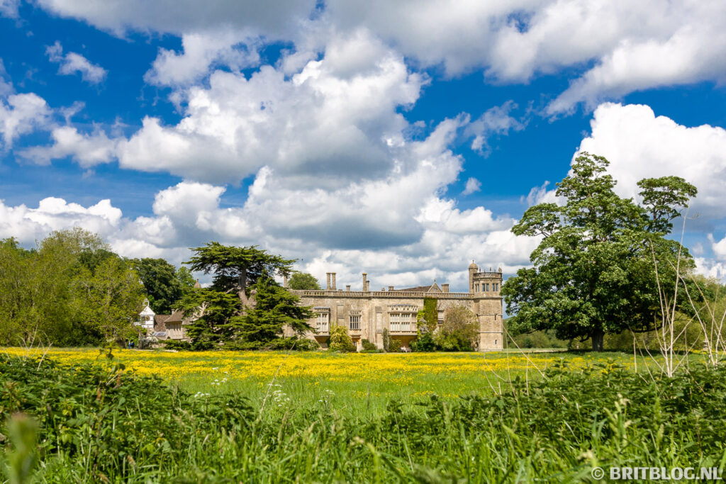 Uitzicht over een bloemenweide op het historische Lacock Abbey in de Cotswolds onder een blauwe lucht met stapelwolken. Dit is een van de vele bezienswaardigheden in de Cotswolds.