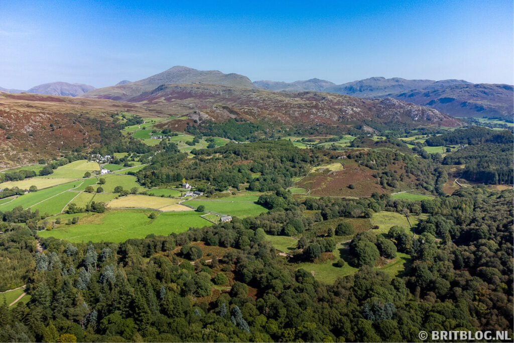 Luchtfoto van de Eskdale-vallei in het Lake District met groene weilanden, heuvels en beboste hellingen onder een heldere blauwe lucht.