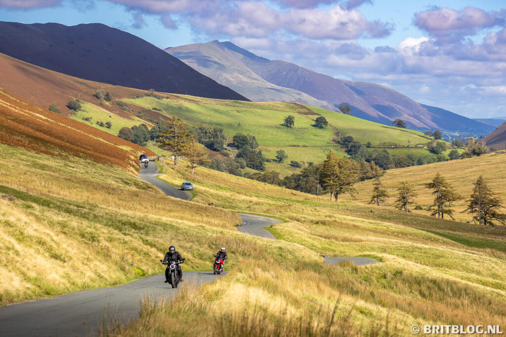 Kronkelende weg door de Newlands Pass in het Lake District met motorrijders, auto’s en groene heuvels onder een blauwe lucht.