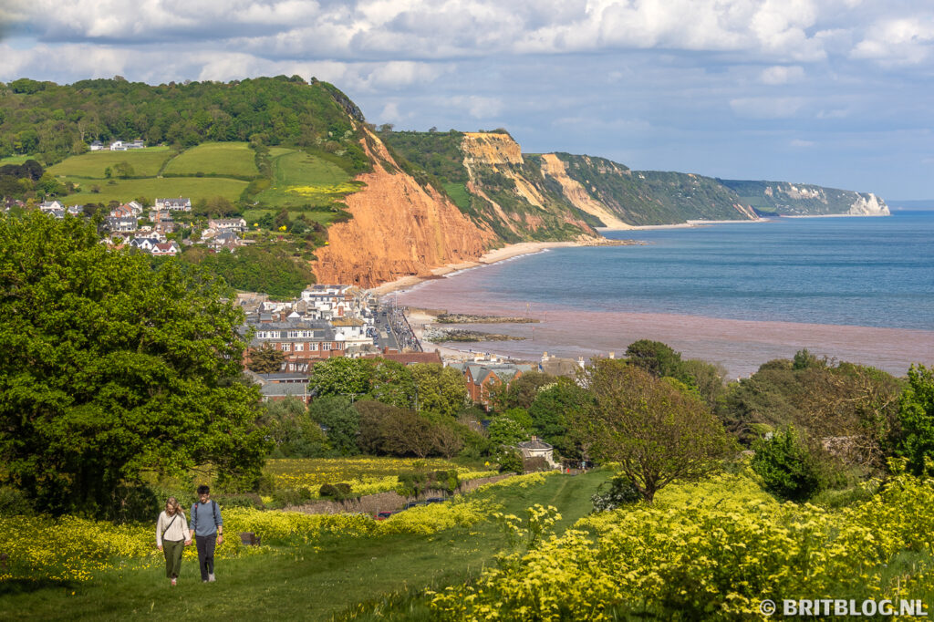 Een paar wandelt over de South West Coast Path naar boven, achter hen zie je Sidmouth, de rode en witte kliffen en de zee.