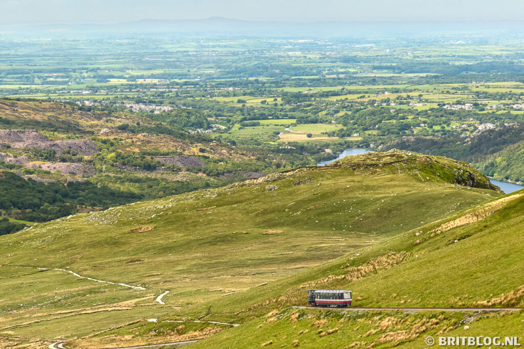 Een treintje rijdt Yr Wyddfa (Mount Snowdon) af, in de verte zie je een meer en lage vlaktes.